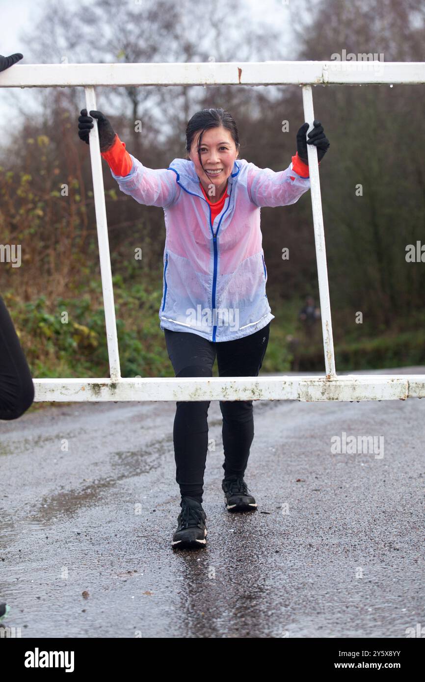 Frau, die einen Hindernislauf im Freien bei nassen Bedingungen absolviert, mit entschlossenem Ausdruck an einer horizontalen Leiter festhält. Stockfoto