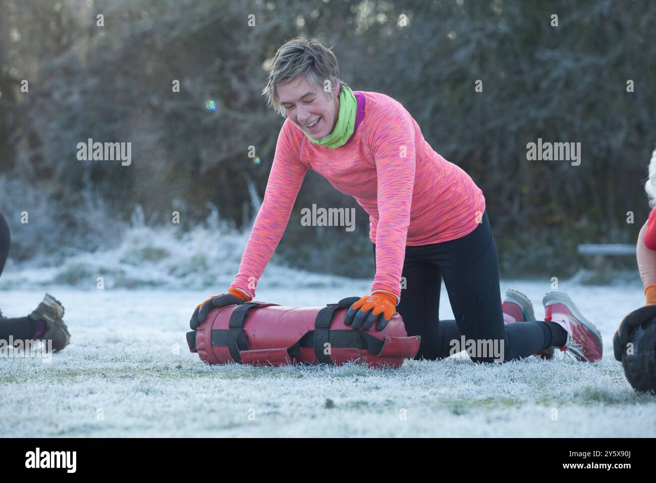 Frau lacht, während sie Outdoor-Übungen mit einem gewichteten Beutel auf einem frostigen Feld macht. Stockfoto