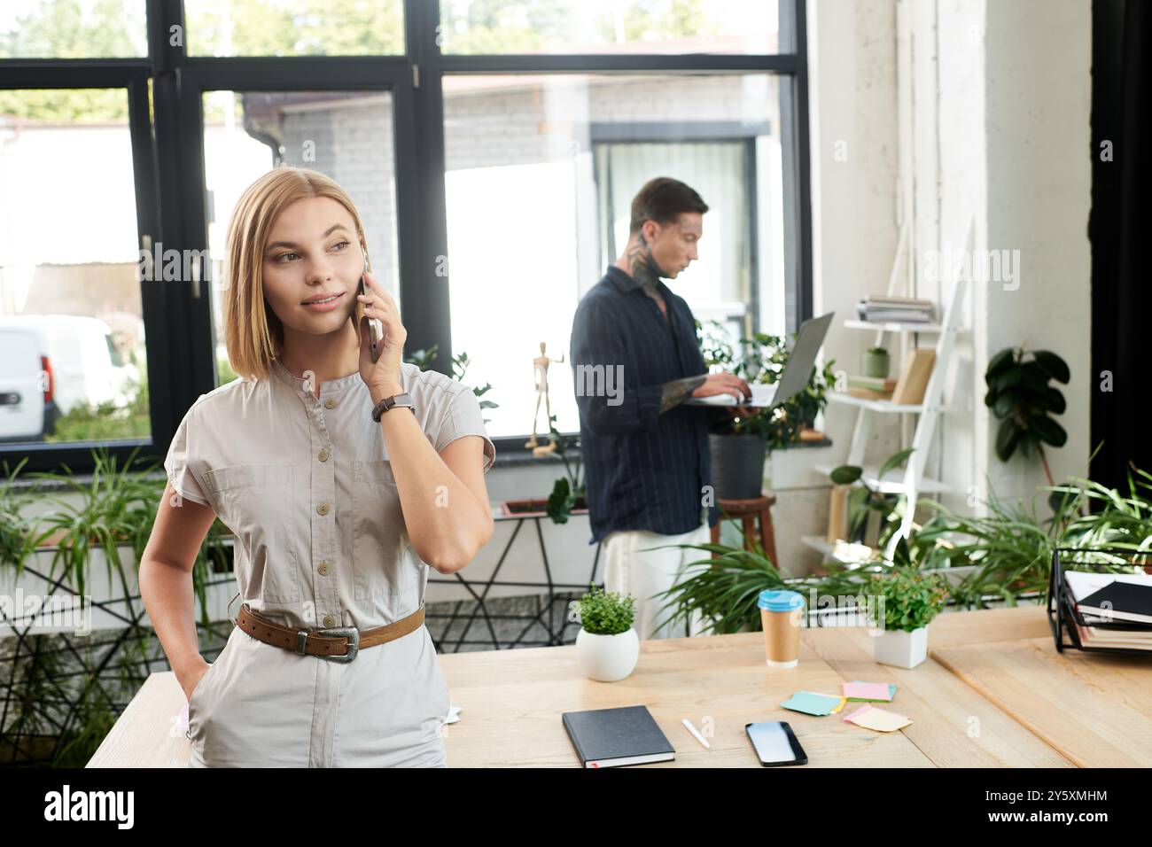 Eine junge Frau spricht am Telefon, während ein Kollege in einem schicken Büro an seinem Laptop arbeitet. Stockfoto