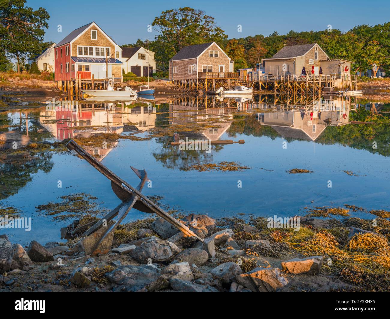 Küstenhäuser spiegeln sich in ruhigem Wasser, mit verankerten Booten und einem alten Anker im Vordergrund. Die Szene spielt vor dem Hintergrund von Bäumen, Creat Stockfoto