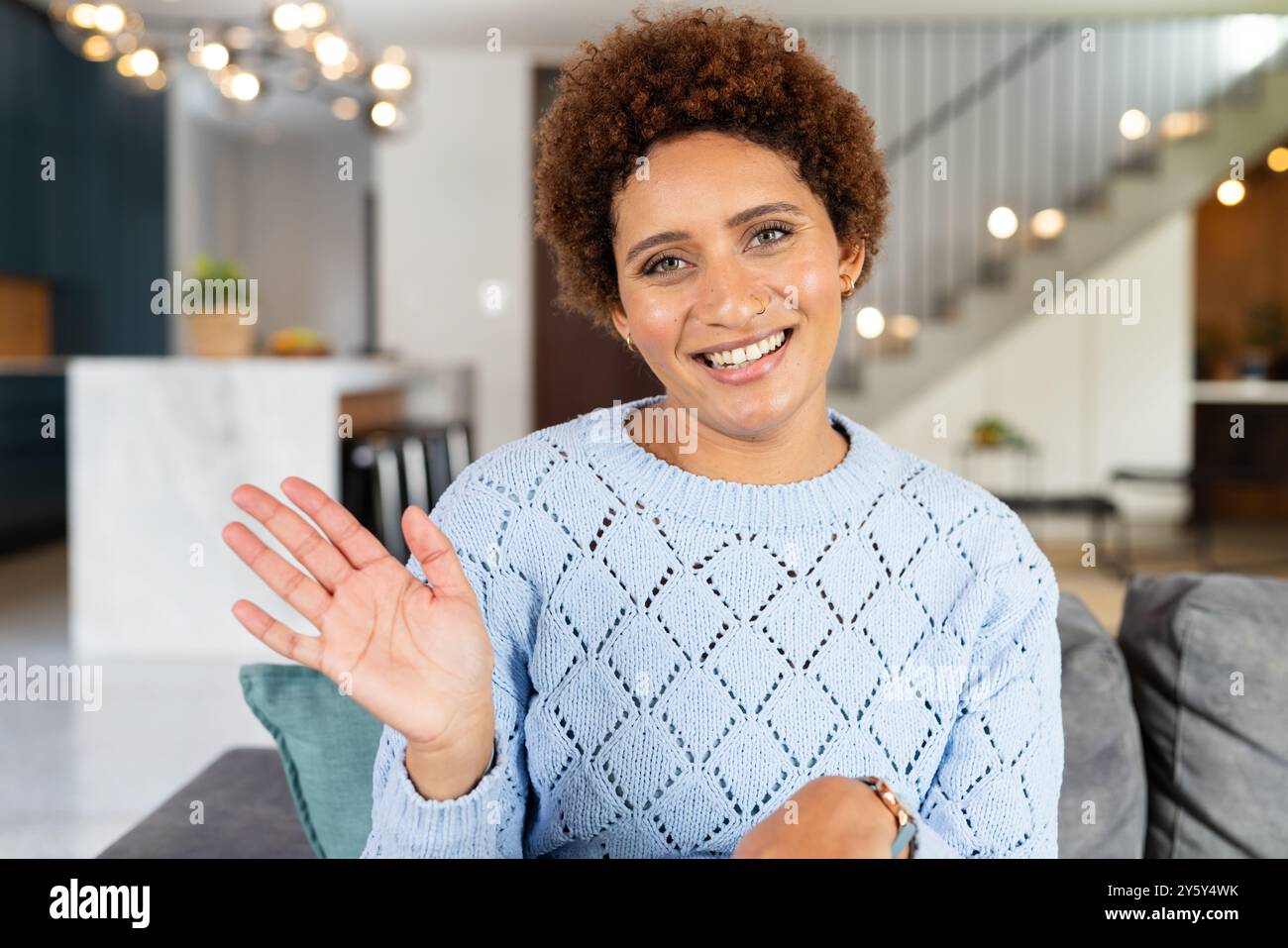 Lächelnde Frau, die Hand winkt, während sie auf der Couch im modernen Wohnzimmer sitzt, auf Videoanruf Stockfoto