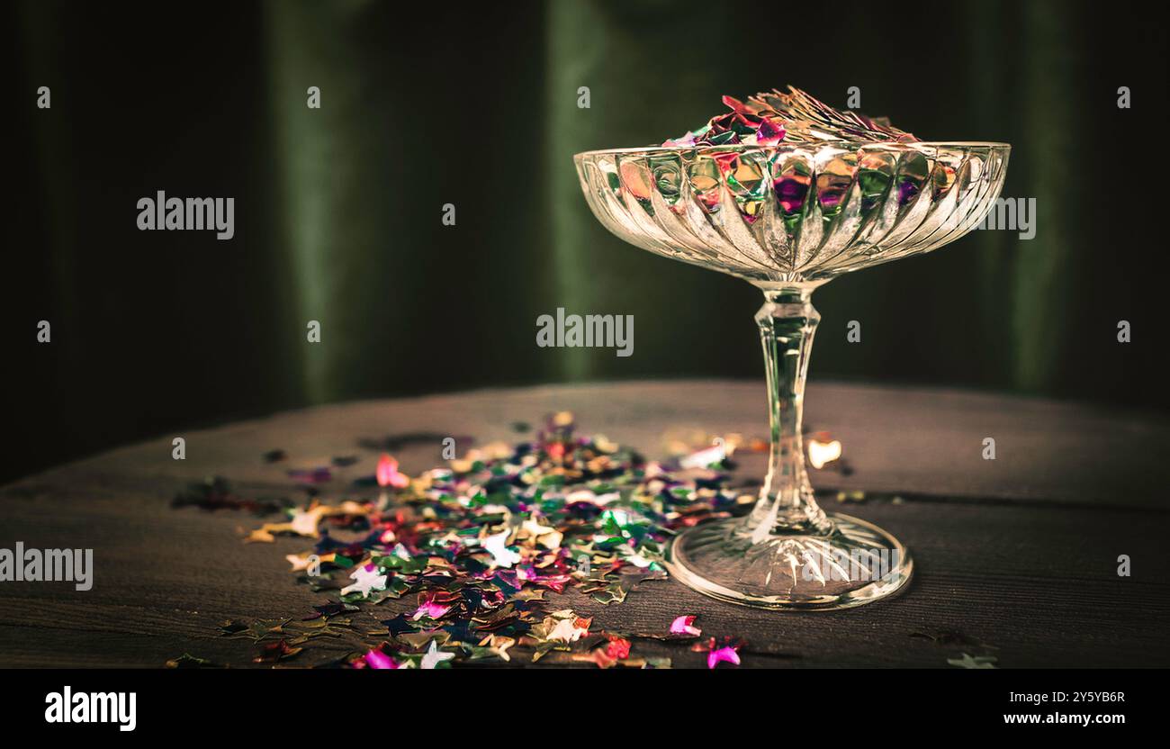 Sektuntertasse im Vintage-Kristallschliff, Glascoupé gefüllt mit farbenfrohem Konfetti, das auf einer Party auf eine Tischplatte geschüttet wird, dunkelgrüner Samthintergrund Stockfoto