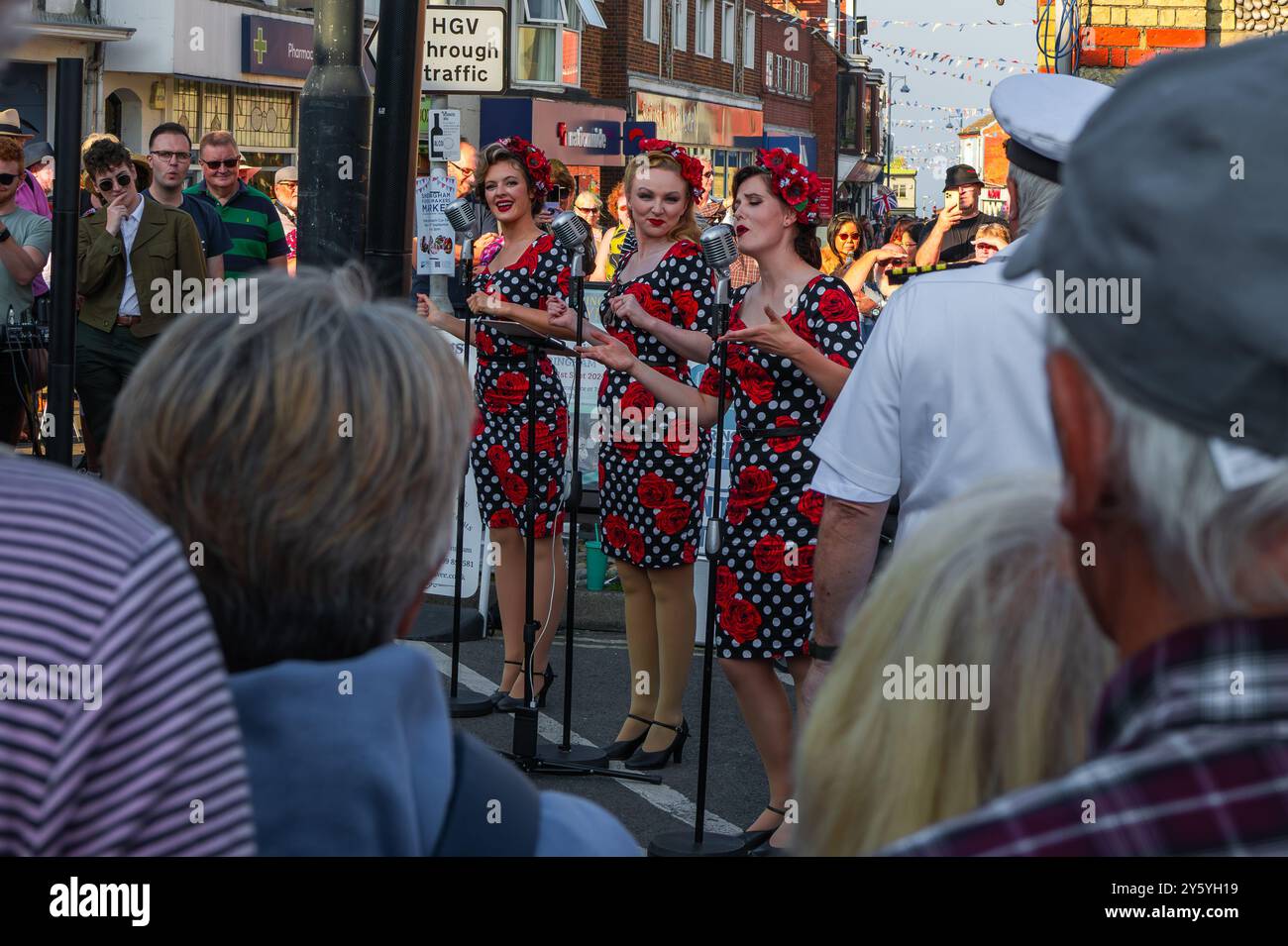 Sheringham, Norfolk, Großbritannien - 21. SEPTEMBER 2024: Foto von drei Frauen in schwarzen Vintage-Kleidern mit weißen Flecken und Rosen, die vor Retro-Microp singen Stockfoto