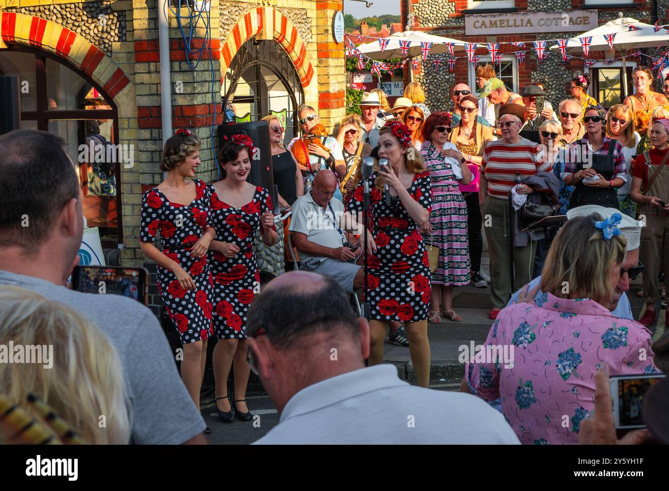 Sheringham, Norfolk, Großbritannien - 21. SEPTEMBER 2024: Foto von drei Frauen in schwarzen Vintage-Kleidern mit weißen Flecken und Rosen, die vor der Menschenmenge singen Stockfoto