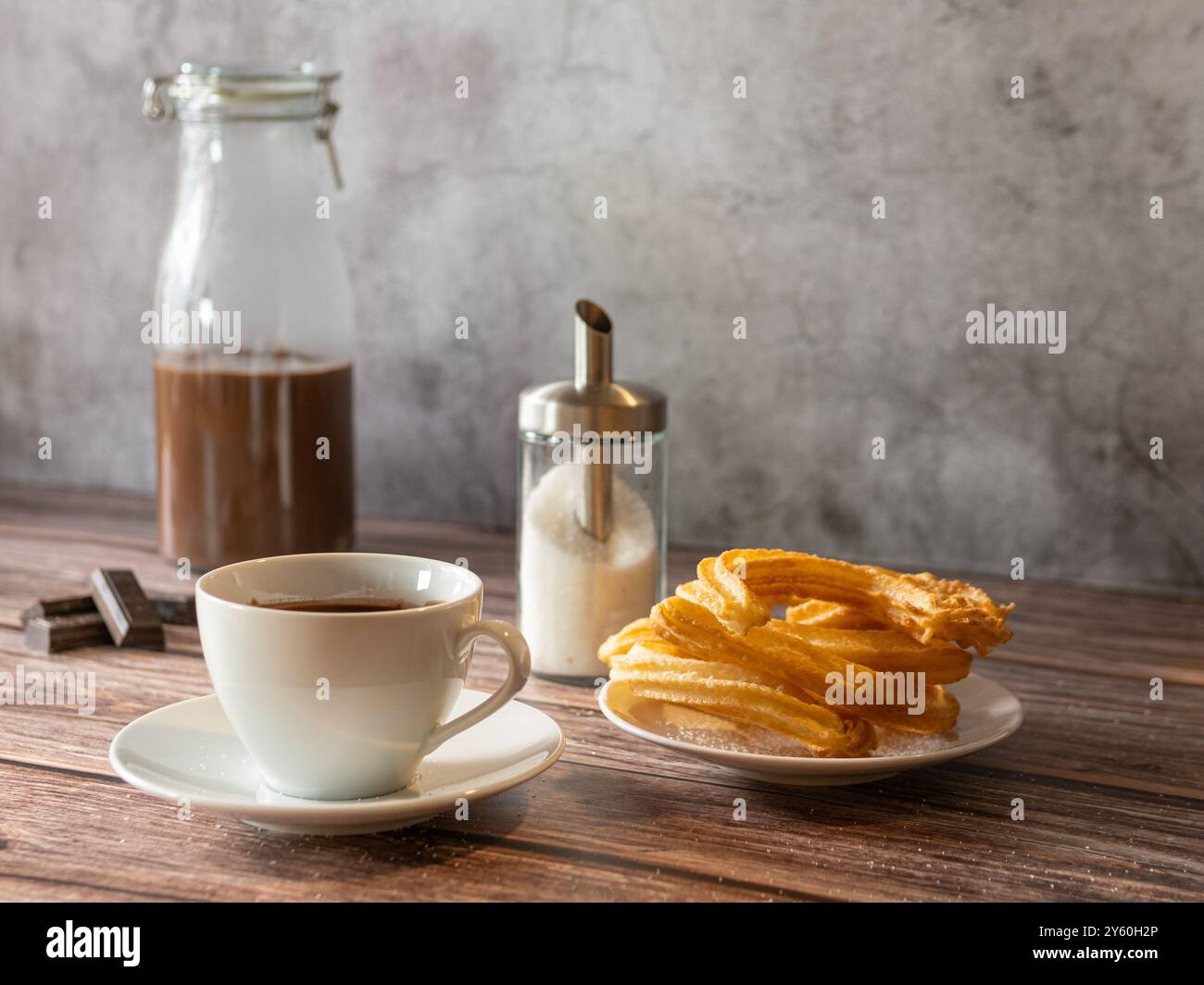 Stillleben einer Tasse heißer Schokolade mit Churros, Glasflasche mit Schokolade am Boden und Glaszuckerschale. Holztisch Stockfoto