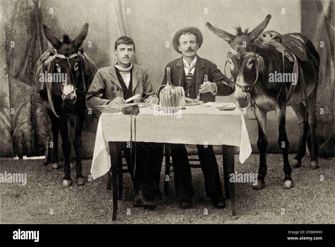 ITALIEN , 1900 c. : kurioses Bild, das draußen auf einem Set aufgenommen wurde, das von einem professionellen Fotografen für diesen Anlass neu gebaut wurde, mit zwei jungen Männern, die an einem Tisch sitzen, um in Begleitung ihrer beiden Esel zu essen. Es ist nicht bekannt, ob dies auf den Verzehr von Pferdefleisch anspielt. Foto gemacht in Norditalien in einer ländlichen Bauernwelt. Unbekannter Fotograf. - NORDITALIEN . - ITALIA - UOMINI - UOMO - DIVERTIMENTO - BUFFO - LUSTIG - FOTO STORICHE - GESCHICHTE - 900ER - '900 - NOVECENTO - XX CENTURY - GIOVENTÙ - JUNG - GIOVANE - MONDO CONTADINO - CONTADINI - LANDWIRTSCHAFT - BAUERNHOF Stockfoto