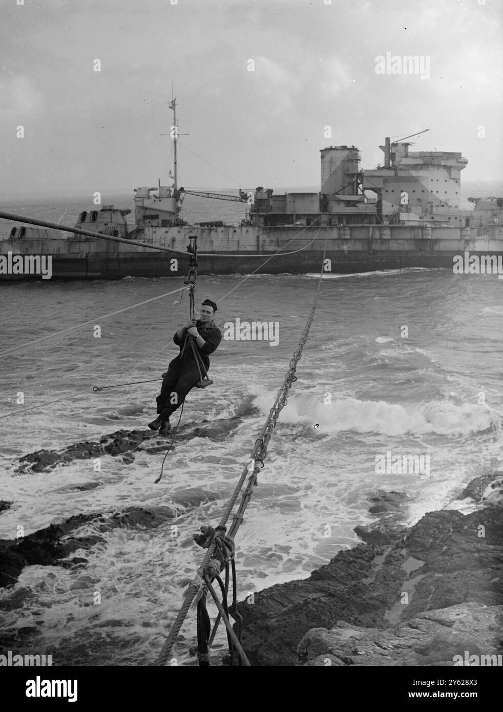 Tag und Nacht arbeiten, um möglichst viel Metallschrott aus dem zerschlagenen Rumpf des tapferen alten Schlachtschiffs „Warspite“ zu retten, das nun in Prerussia Cove in Cornwall zerbrochen wurde, und die Arbeiter benutzen eine Seilbahn, um bei rauem Wetter zum und vom Schiff zu fahren. Die Bergung wurde in der letzten Woche beschleunigt, hat einen Riss in der Schiffsstruktur, direkt hinter der Brücke wurde festgestellt. Das Bild zeigt: Hoch über den wütenden Wellen kommt ein Arbeiter aus der „Warspite“ über die Seilbahn „Bosun's Sessel“. 16. Januar 1948 Stockfoto
