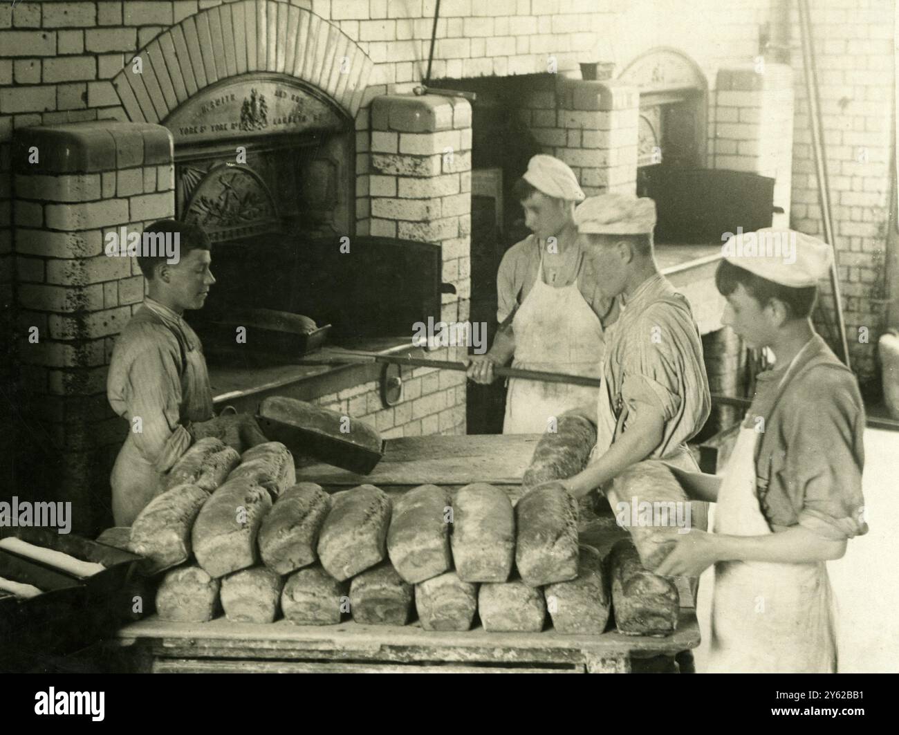 ARCHIV BOX 4 ARC 0129 Referenz: 001415 THE BAKERY : BOYS GARDEN CITY , Woodford Bridge , Essex , England DATUM : 1920ER JAHRE JUNGEN, DIE GEBACKENE BROTE AUS DEM OFEN NEHMEN . BROTHAUFEN IM VORDERGRUND. BÄCKER Stockfoto