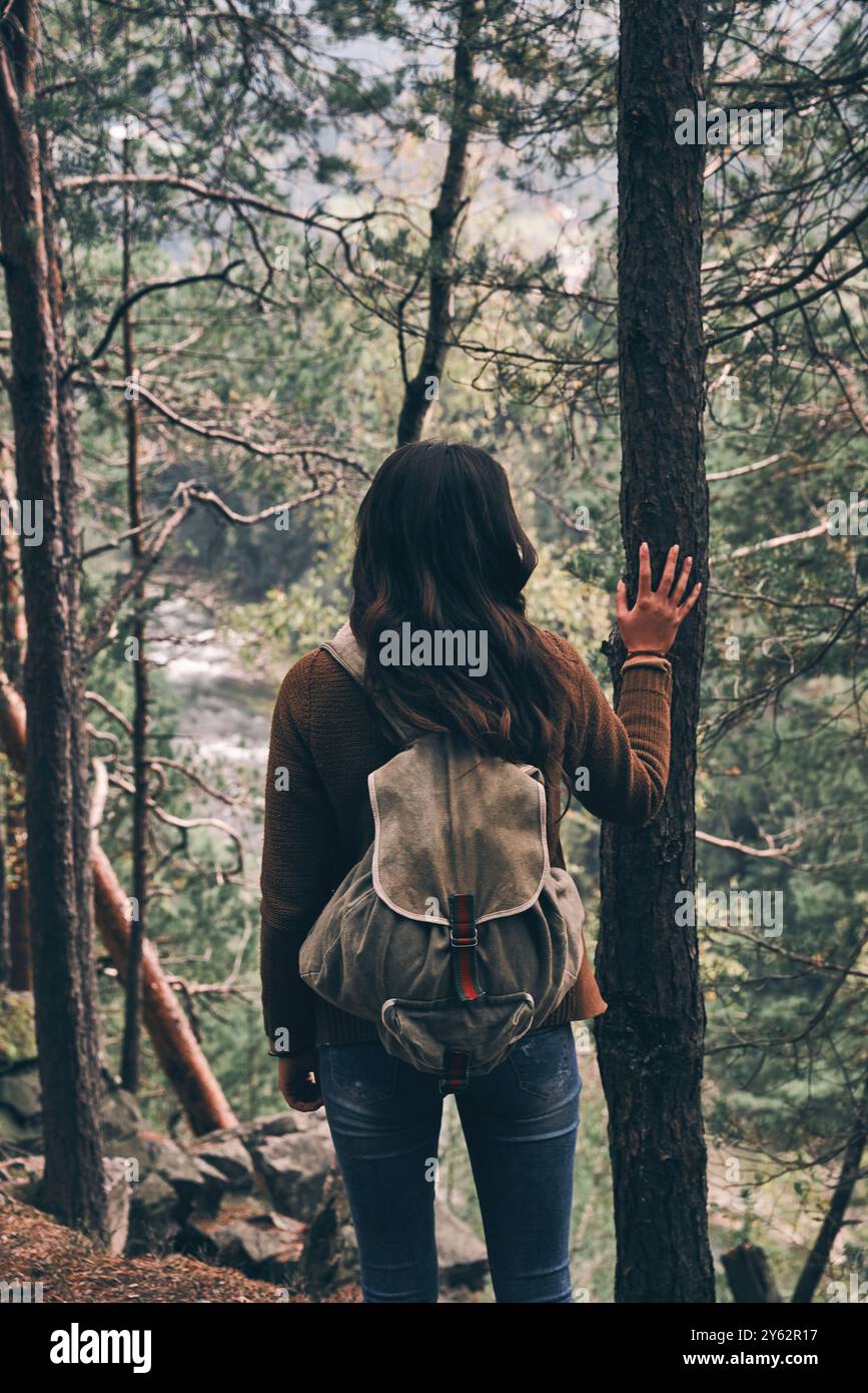 Die Natur fühlen. Rückansicht einer jungen modernen Frau mit Rucksack, die den Baum berührt und die Aussicht bewundert Stockfoto