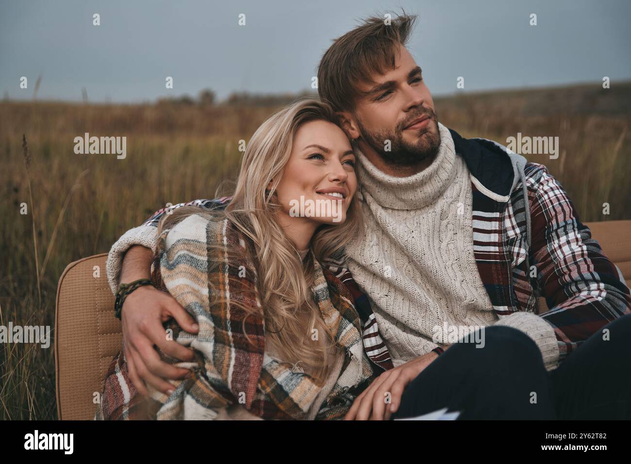 All die Sorgen sind weggeflogen... Wunderschönes junges Paar, das auf dem Feld sitzt und mit einem Lächeln wegblickt Stockfoto