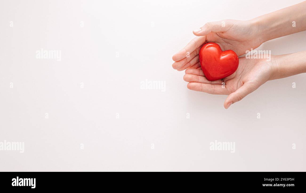 Ein Paar Hände, die sanft ein rotes Herz wiegen und Liebe, Mitgefühl, Sorgfalt und Freundlichkeit vor einem weichen, neutralen Hintergrund darstellen Stockfoto