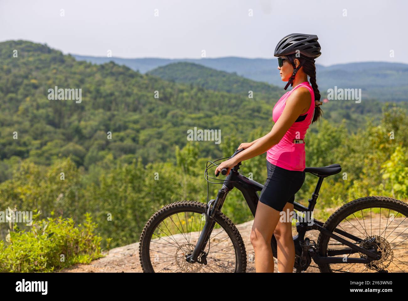 Frau auf E-Mountainbike in rosa Spitze, Helm und Sonnenbrille, genießen die malerische Aussicht auf den Wald. EMTB-Konzept Stockfoto