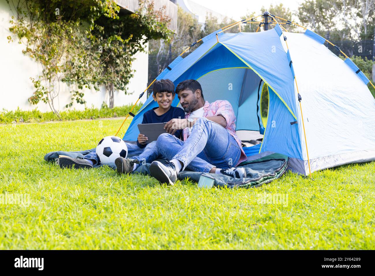 Camping im Hinterhof, indischer Vater und Sohn mit Tablette und Entspannung in der Nähe des Zeltes Stockfoto