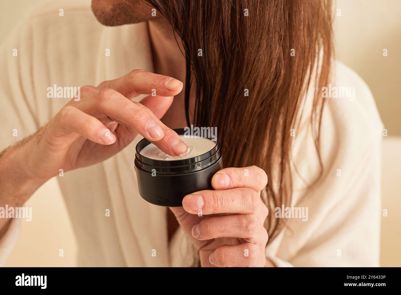 Mann mit langen Haaren nimmt weiße Creme aus schwarzem Glas mit Finger-Nahaufnahme, tägliche Körperpflege für Männer Stockfoto