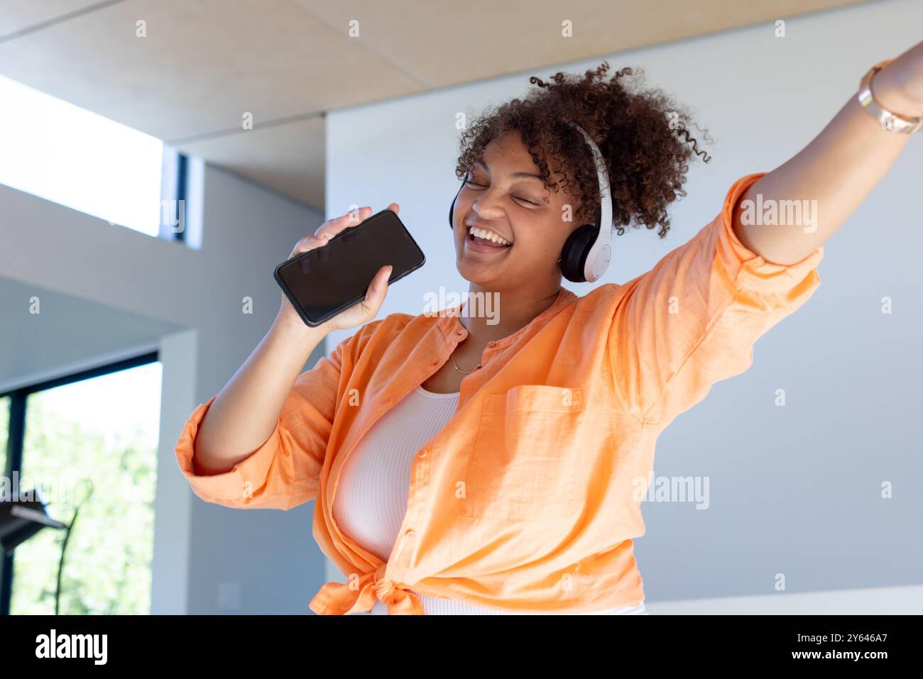 Sie trägt Kopfhörer und eine Frau in Größe, die zu Hause singt und ihr Smartphone hält Stockfoto