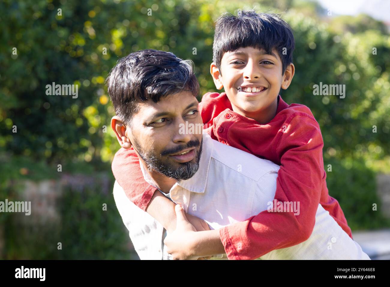 Ein indischer Vater reitet dem jungen Sohn mit Huckepack, beide lächeln draußen Stockfoto