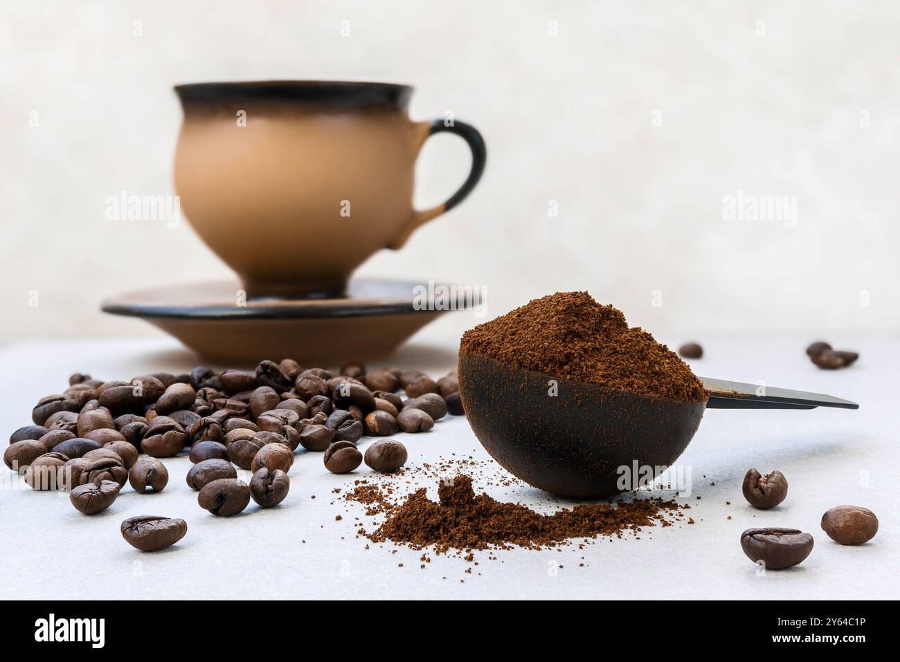 Frisch gemahlener aromatischer Kaffee in einem Messlöffel vor einem Hintergrund von verstreuten Kaffeebohnen und einer Keramikbecher und Untertasse im Hintergrund Stockfoto