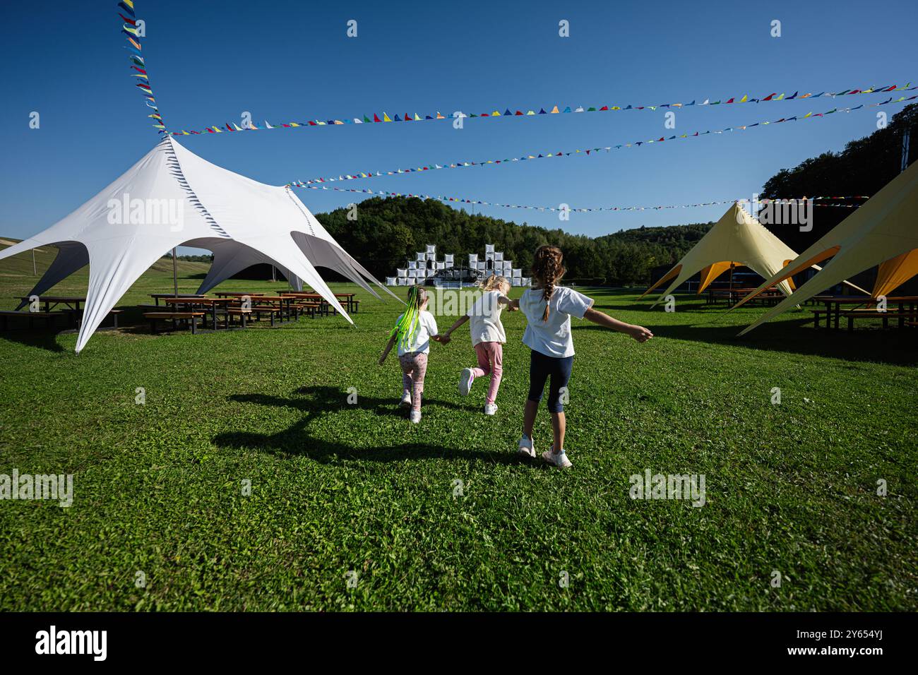 Drei fröhliche Kinder laufen bei einem Outdoor-Festival durch ein grasbewachsenes Feld. Helle Zelte und Fahnen sorgen für eine festliche Atmosphäre unter einem klaren blauen Himmel. Stockfoto