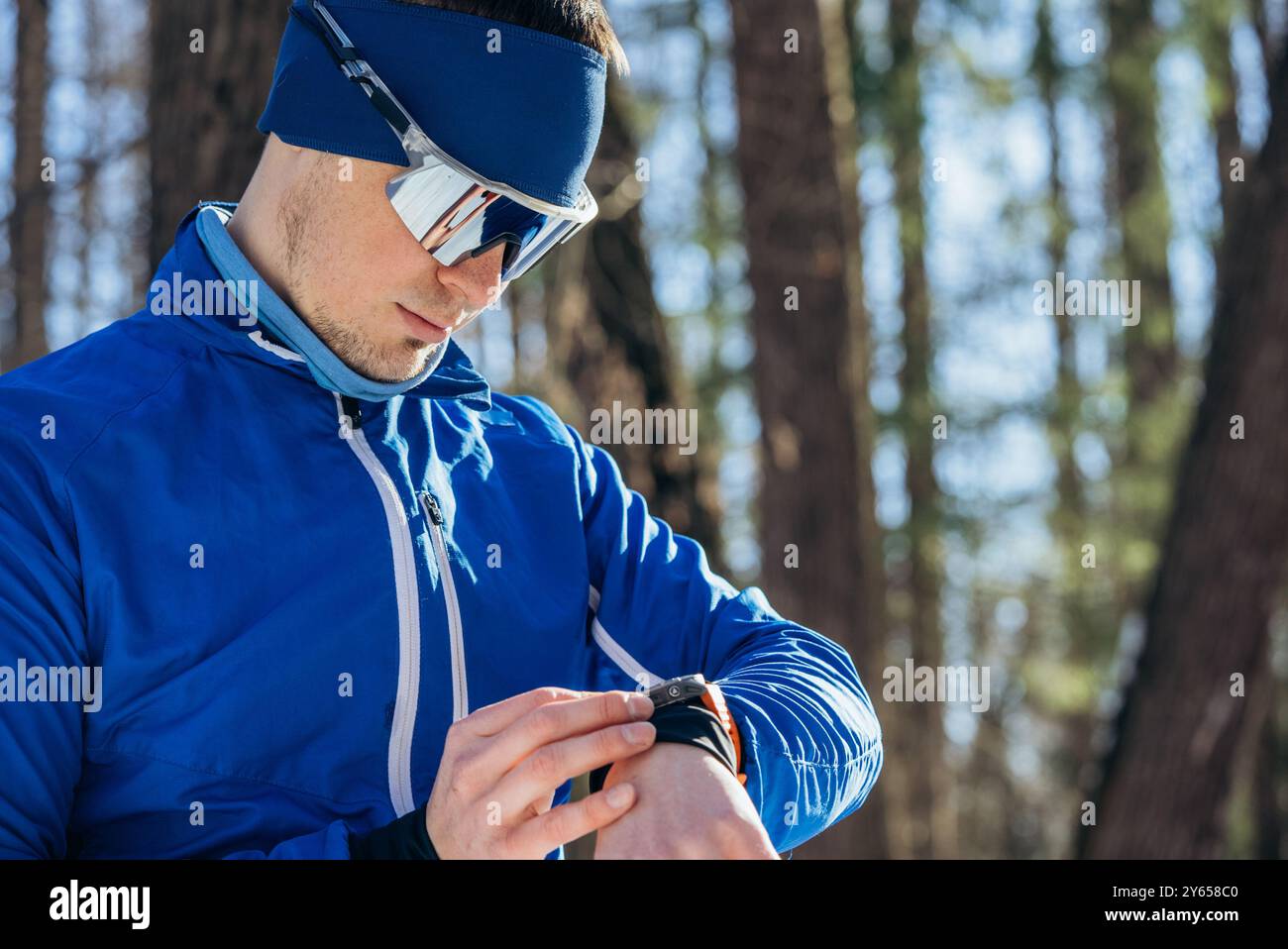 Sportlicher Mann in Wintersportbekleidung, der Smartwatch beim Workout im Wald verwendet, kaltes Wetter-Training. Er trägt eine reflektierende Sonnenbrille und ein Stirnband. Stockfoto