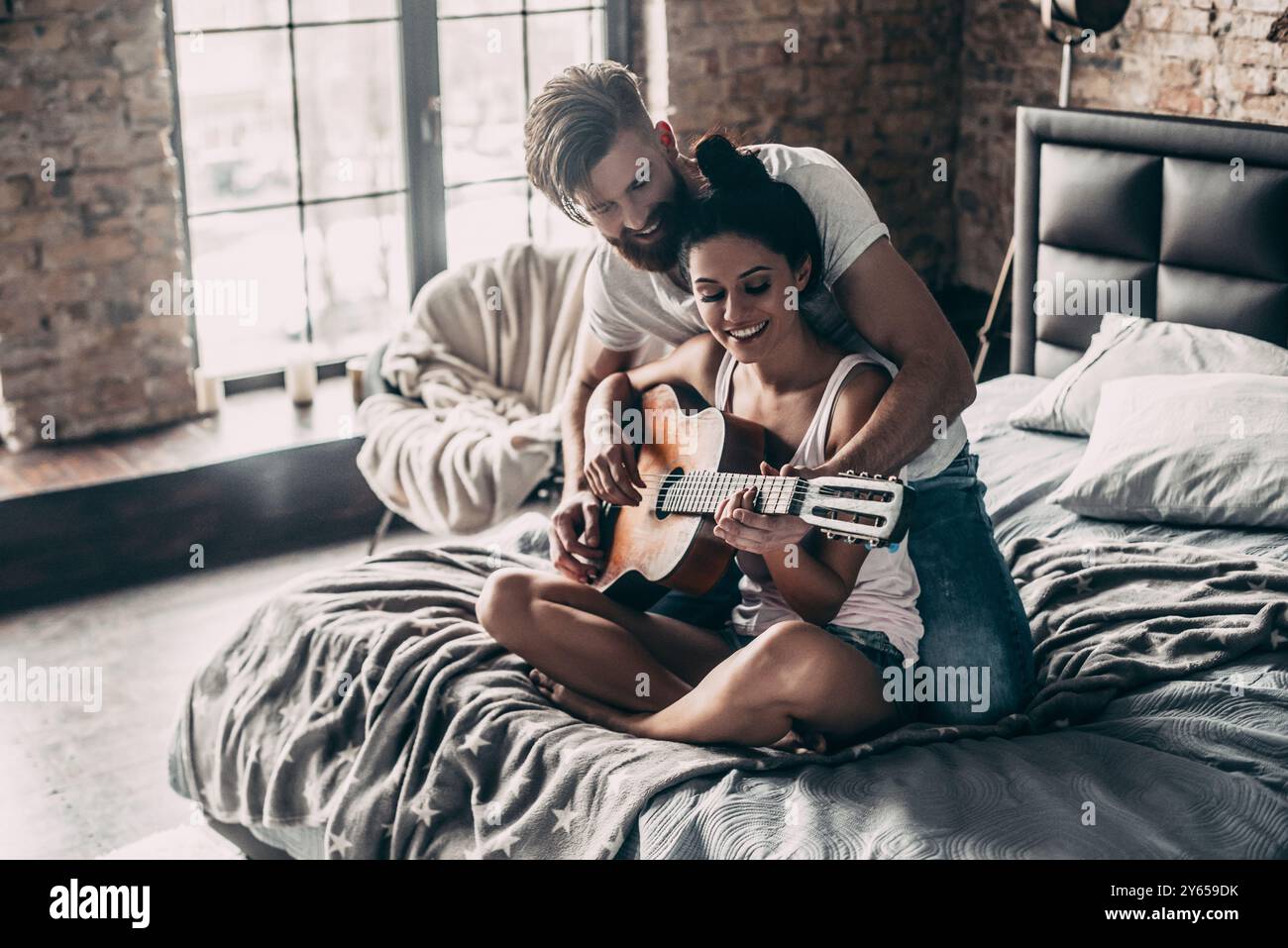 Du spielst großartig! Hübscher junger bärtiger Mann, der seiner Freundin beibringt, Gitarre zu spielen, während beide zusammen im Bett sitzen Stockfoto