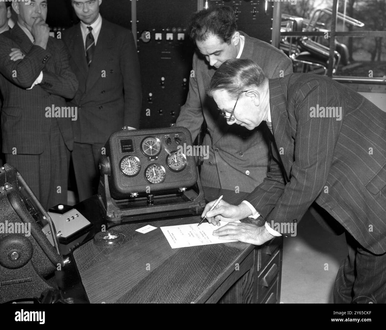 In einer Funkstation in Puckeridge , Herts , hat der Abgeordnete von Rt Hon Alfred Barnes , Verkehrsminister , offiziell den Decca Navigator Identification Service ins Leben gerufen . Eine unschätzbare Innovation für den Luft - und Seeverkehr , dieses System von Funkspuren , die von der Station aus zu einem Gitterwerk von Wellen ausgesandt wurden , werden auf zwei Decometern aufgezeichnet , mit denen das Schiff seine Position innerhalb von zehn Metern in einer beträchtlichen Entfernung von der Station bestimmen kann , Foto zeigt : nach der Inbetriebnahme des Systems wird Barnes gesehen , wie er das erste Logbuch der Station signiert . Links ist der Erfinder Mr. Wi Stockfoto