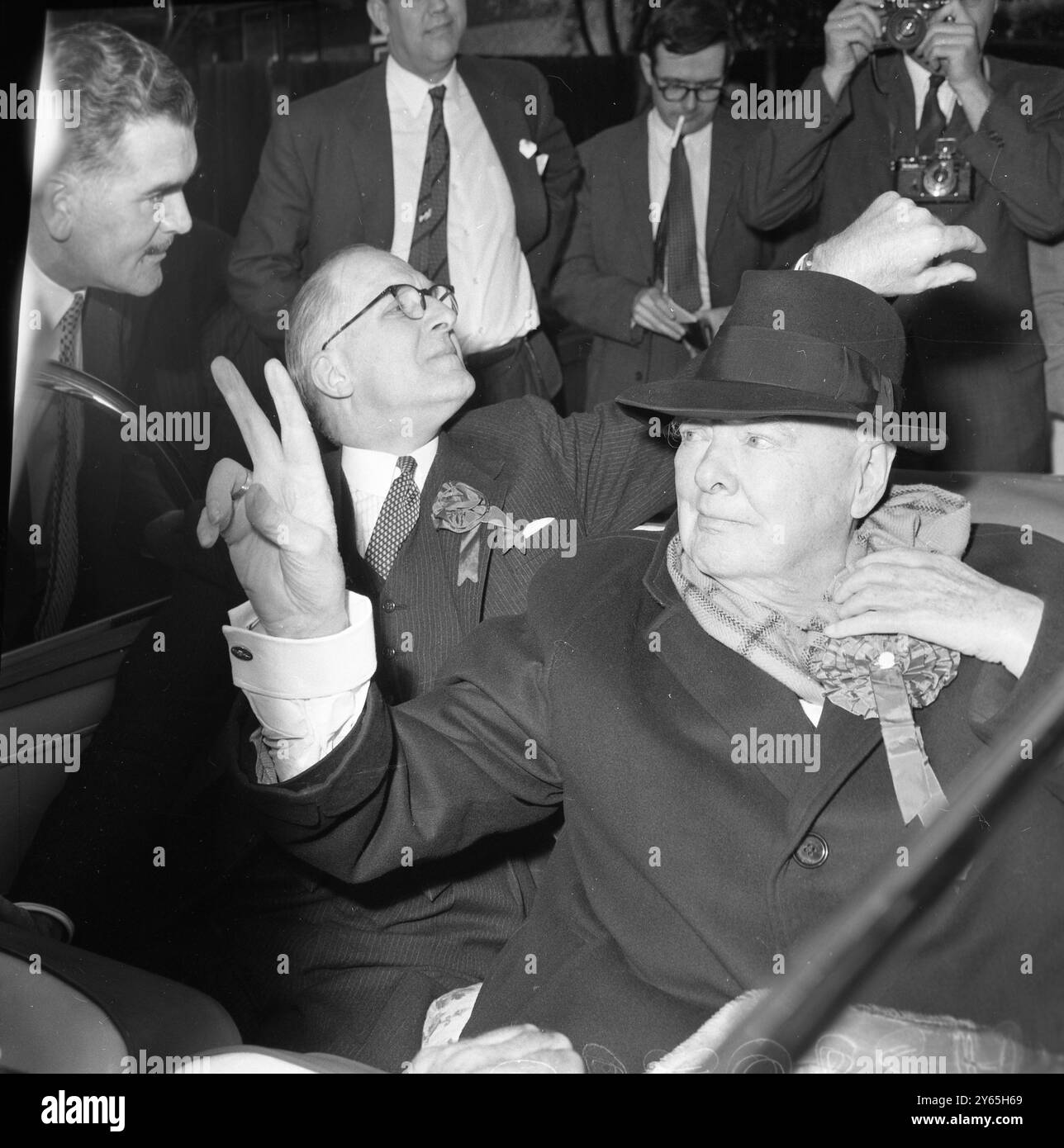 Oktober Heat der 84-jährige Ex-Premierminister Sir Winston Chyrchill zieht die Krempe seines Hutes nach unten , um seine Augen vor der Sonne zu schützen , zieht seinen Schal während seiner Wahlreise in Woodford heraus und gibt seinen Wählern sein berühmtes V-Zeichen . Oktober 1959 Stockfoto