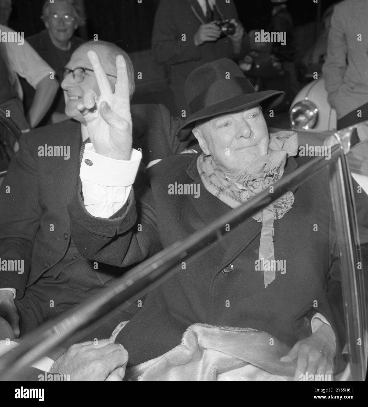 Oktober Heat der 84-jährige Ex-Premierminister Sir Winston Chyrchill zieht die Krempe seines Hutes nach unten , um seine Augen vor der Sonne zu schützen , zieht seinen Schal während seiner Wahlreise in Woodford heraus und gibt seinen Wählern sein berühmtes V-Zeichen . Oktober 1959 Stockfoto