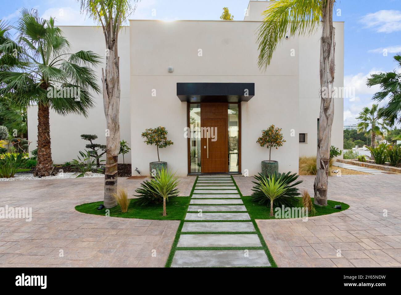 Ein moderner Villeneingang mit einem symmetrischen Gartenweg, umgeben von üppigen Palmen und dekorativen Pflanzen, bietet einen Luxus und in sich Stockfoto
