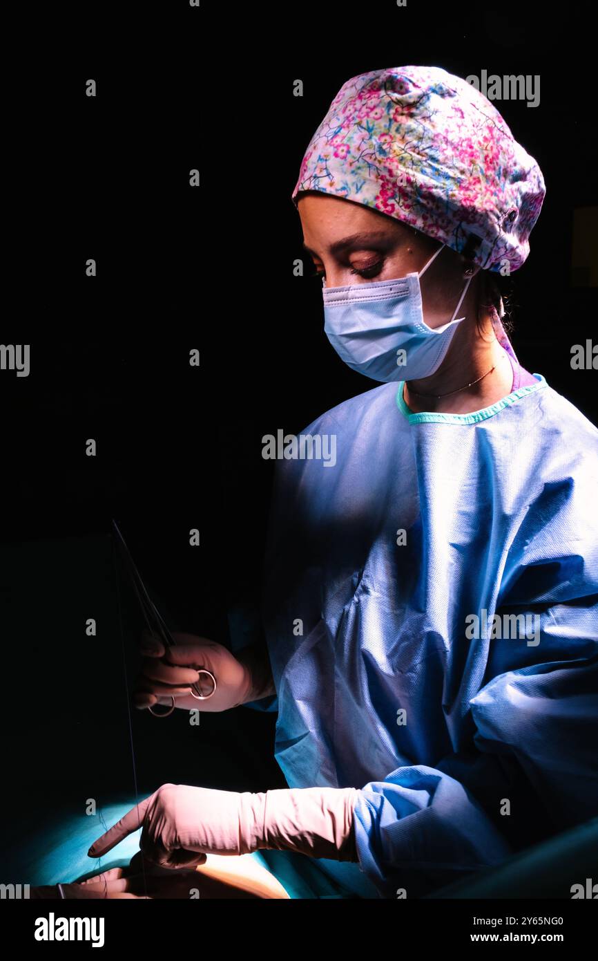 Eine Chirurgin ist während eines chirurgischen Eingriffs intensiv fokussiert und trägt blaue Peelings, eine Blumenkappe und eine blaue chirurgische Maske unter DRAM Stockfoto