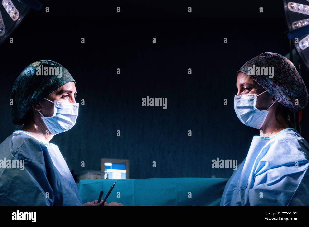 Zwei weibliche Chirurgen stehen in einem schwach beleuchteten Operationssaal von Angesicht zu Angesicht und besprechen die Strategie vor einer Operation. Beide Profis sind in Peelings und tragen s Stockfoto