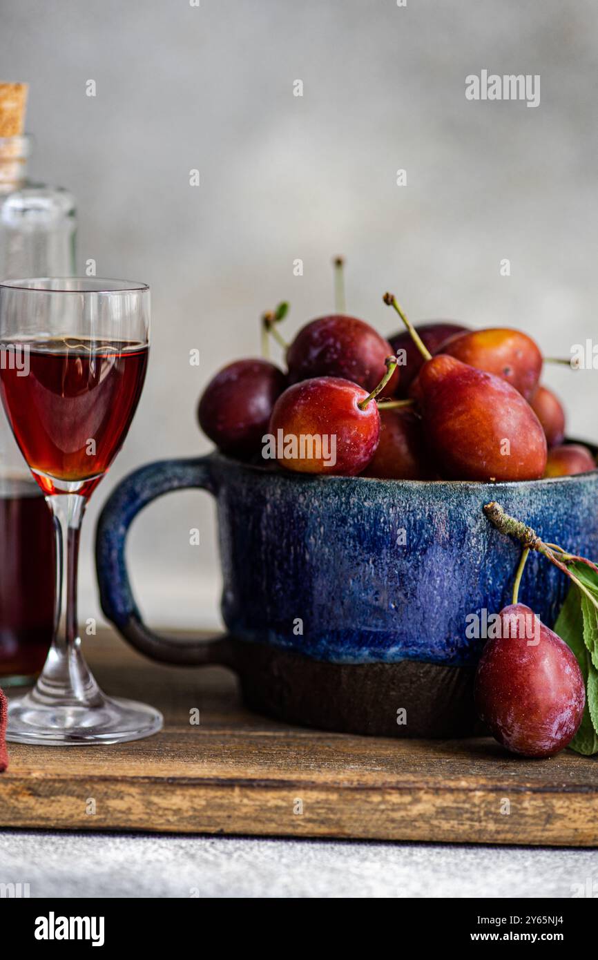 Frische Bio-Pflaumen in einer Keramikschale und ein Glas reichhaltigen Pflaumenlikör, wunderschön auf einer Holzoberfläche ausgestellt. Hausgemachte Authentizität in einem rustikalen K Stockfoto