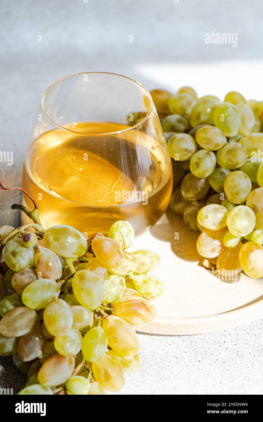 In einer hellen, sonnendurchfluteten Umgebung paart sich ein Glas Weißwein mit frischen Trauben und wirft einen weichen Schatten auf eine strukturierte Oberfläche. Diese einladende sce Stockfoto