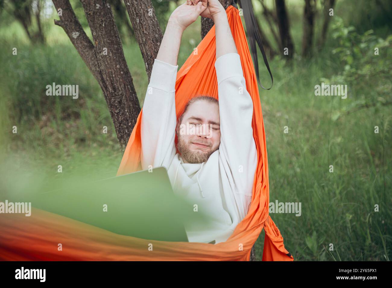 Ein zufriedener junger Mann dehnt sich entspannt in einer leuchtend orangen Hängematte zwischen Bäumen aus, ein Laptop an seiner Seite und verbindet Arbeit und Spiel in einer ruhigen Atmosphäre Stockfoto