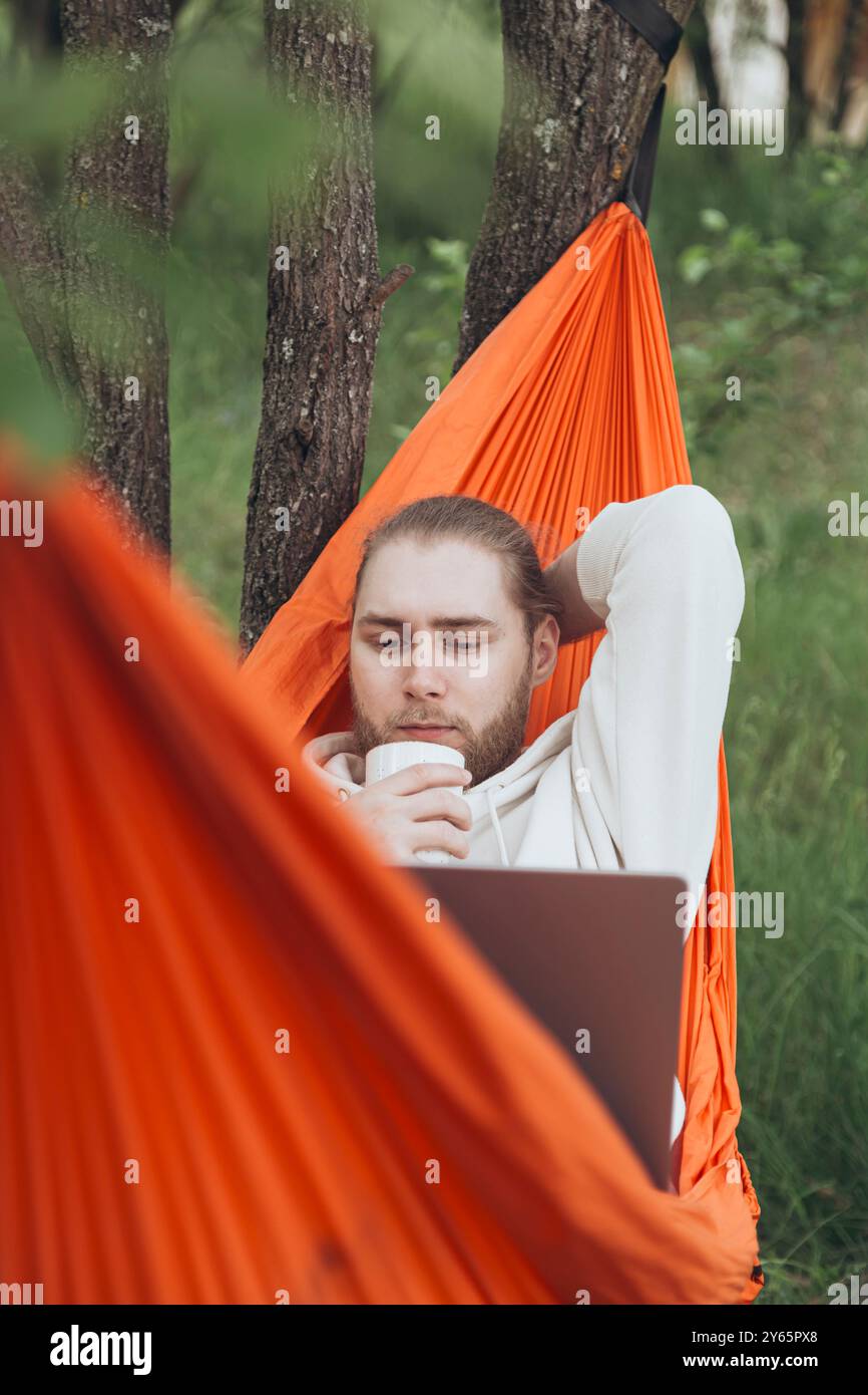 Ein entspannter junger Mann trinkt Kaffee, während er an einem Laptop arbeitet und in einer orangen Hängematte zwischen grünen Bäumen sitzt Stockfoto