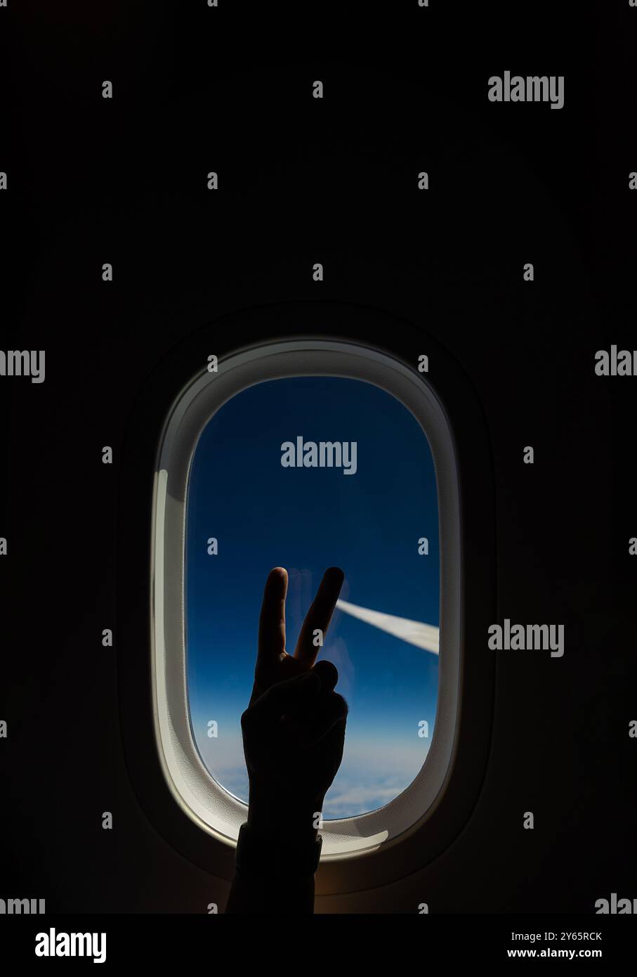 Die Silhouette einer abgeschnittenen, nicht erkennbaren Person, die mit ihren Fingern vor dem Hintergrund eines Flugzeugfensters ein Friedenszeichen macht, das die Reise-sp. Veranschaulicht Stockfoto