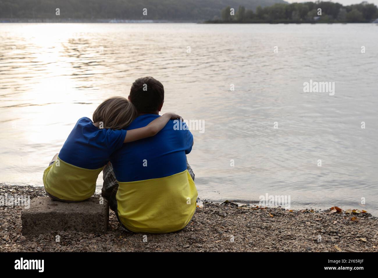 Vater und Sohn sitzen nebeneinander in den gleichen gelben und blauen T-Shirts in den Farben der ukrainischen Flagge am Ufer des Sees Stockfoto