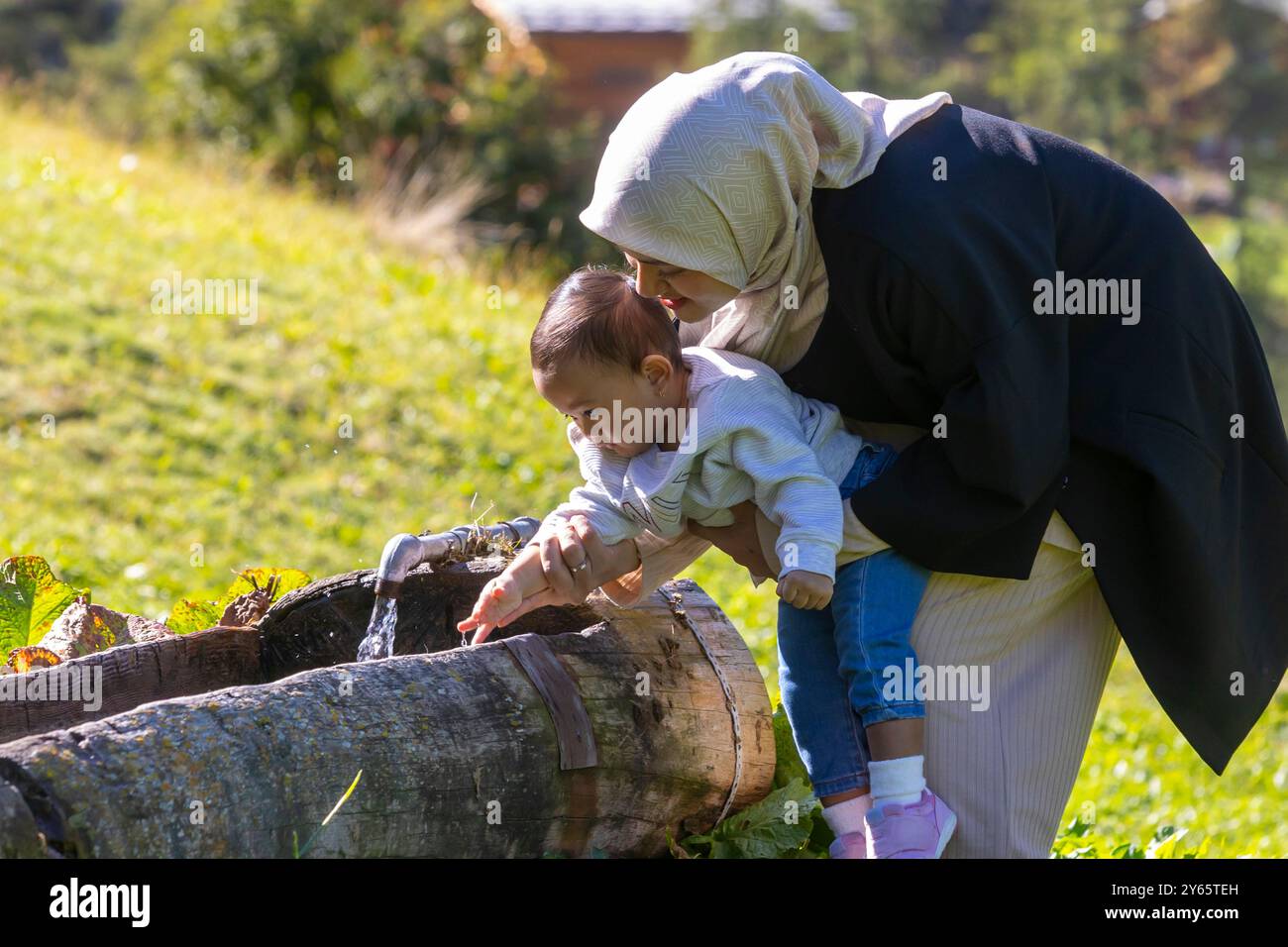 In den Schweizer Bergen entfaltet sich eine berührende Szene, in der eine Mutter in einem Hijab ihrem kleinen Kind sanft mit Wasser interagiert, das aus einem Rusti fließt Stockfoto