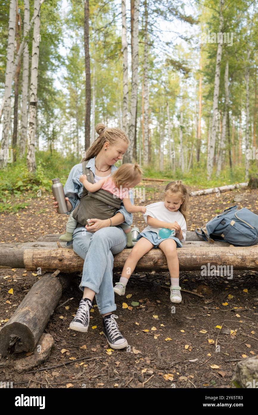 Eine Mutter mit zwei Töchtern, die auf einem Baumstamm im Wald sitzen und einen Moment der Freude während eines Wanderabenteuers teilen, umgeben von einem Birkenwald Stockfoto