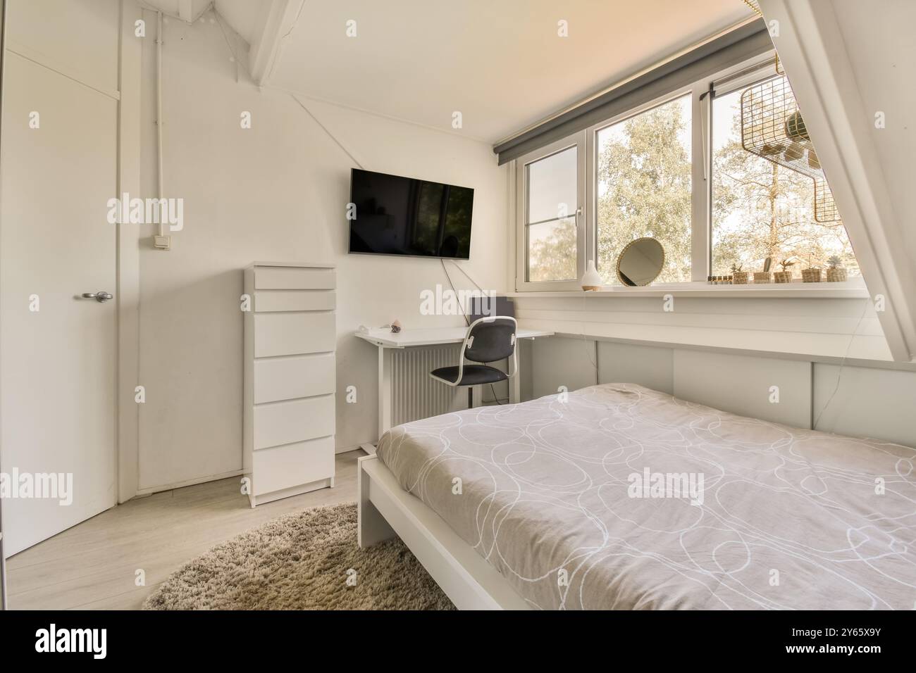 Ein gut beleuchtetes, minimalistisches Schlafzimmer mit Einzelbett, flauschigem Teppich, elegantem Schreibtisch und Flachbildfernseher an der Wand. Stockfoto