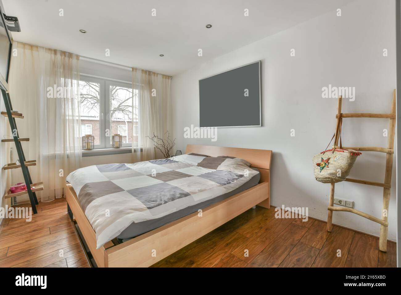 Modernes Schlafzimmer mit einem großen Bett mit karierter Federdecke, Hartholzböden, transparenten Vorhängen und schwarz gerahmten Wandbildern. Stockfoto