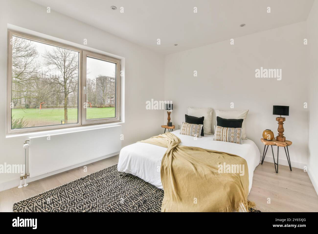 Ein gemütliches und stilvolles, modernes Schlafzimmer mit großem Fenster und Panoramablick auf den Park, dekoriert mit dekorativen Kissen und einer goldenen Decke. Stockfoto