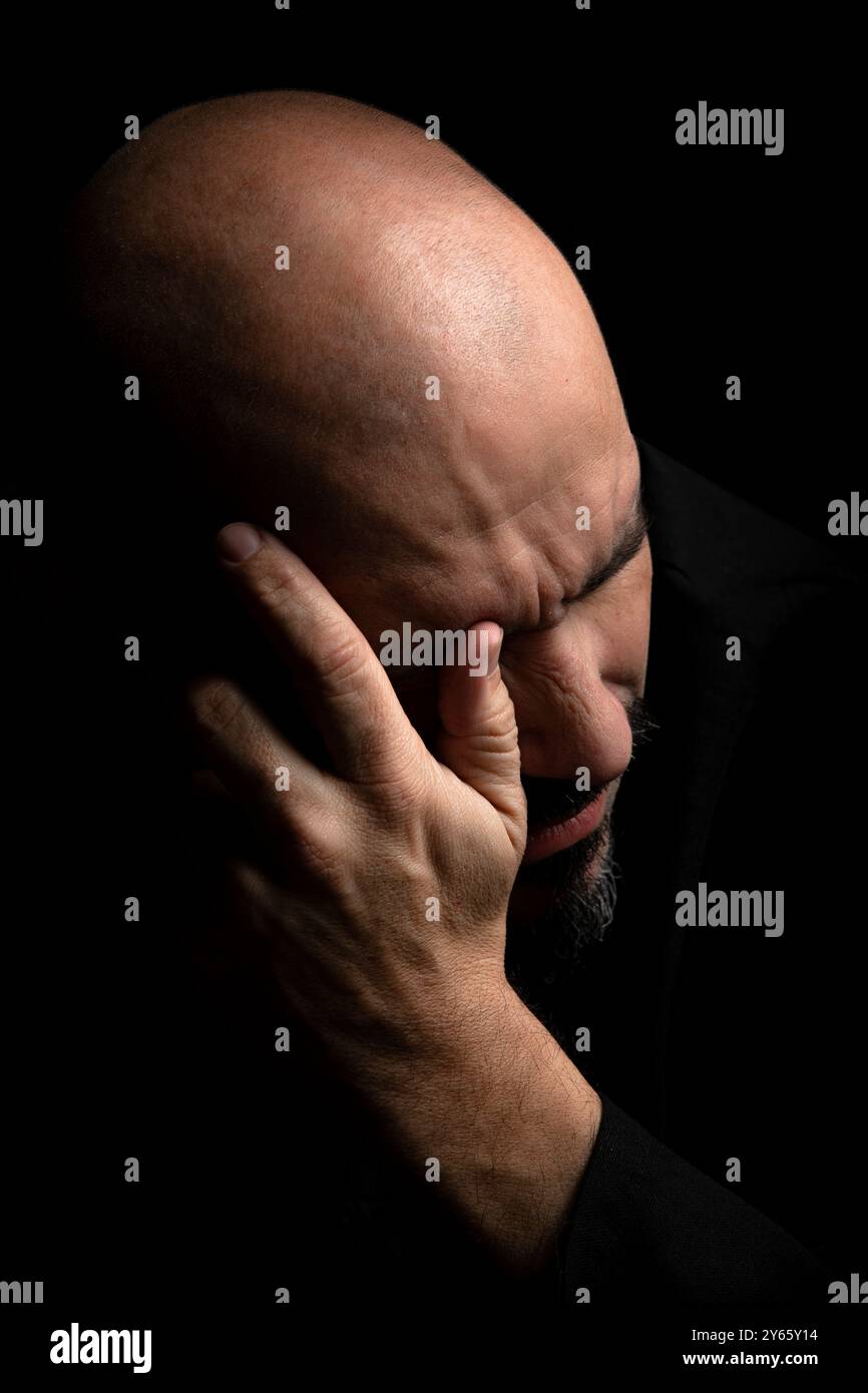 Eine Nahaufnahme eines verzweifelten Glatzen-Mannes, der einen Teil seines Gesichts und seiner Stirn mit seiner Hand bedeckt. Er scheint in Trauer oder Schmerz zu sein, gegen einen dunklen, schwarzen Mann gestellt Stockfoto