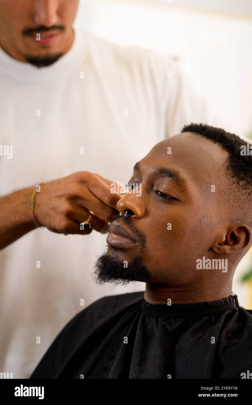 Ein schwarzer Friseur formt sorgfältig die Koteletten eines schwarzen Mannes in einer entspannten Küsten-Barbershop-Umgebung und hebt die spezielle Pflege hervor Stockfoto