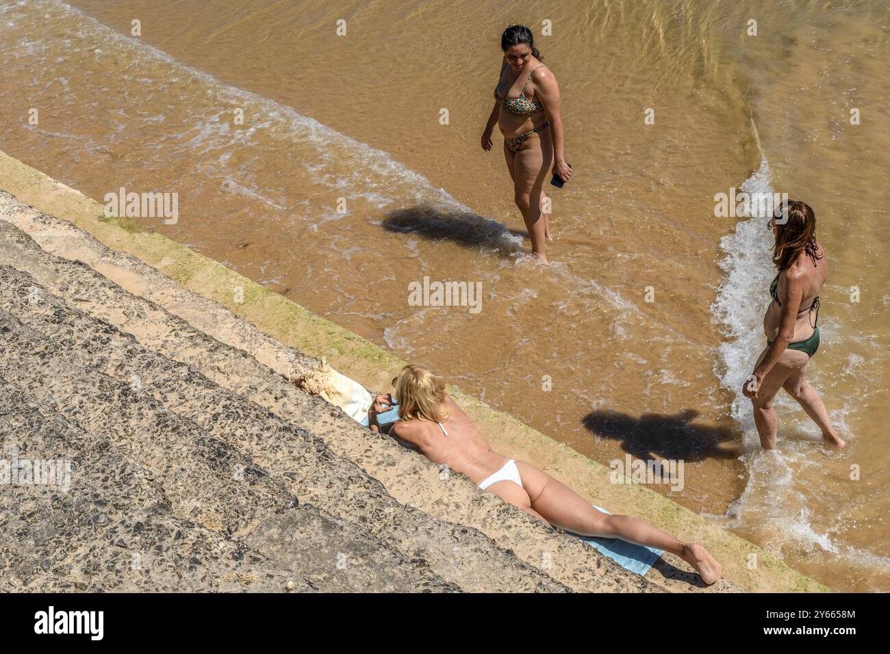 Drei Frauen in Bikinis, eine liegend auf den Stufen des Alberto Romano Ozeanpools in Cascais und zwei mit den Füßen im Wasser. Stockfoto