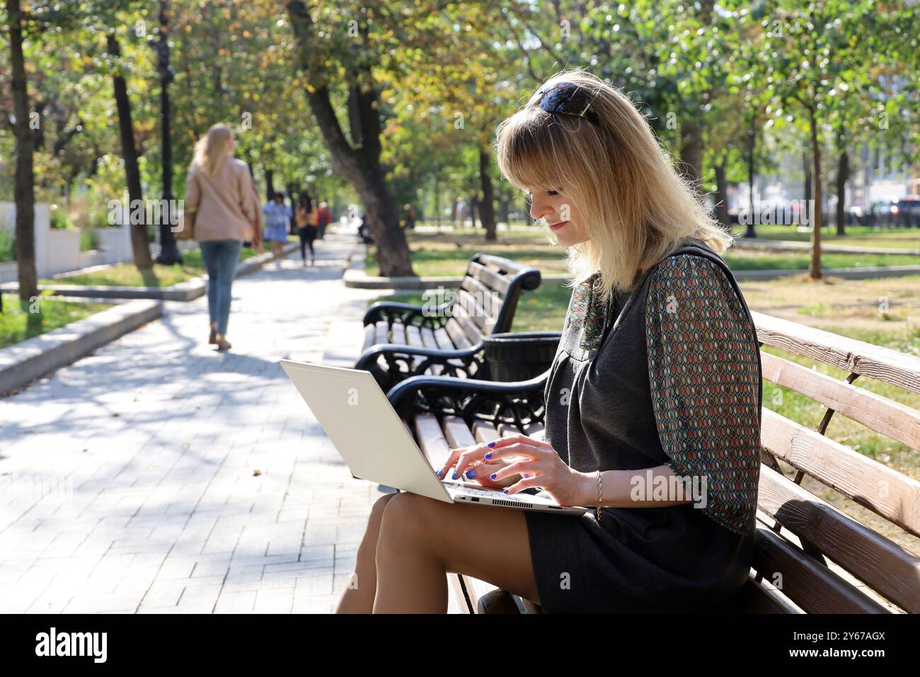 Blonde Frau sitzt mit Laptop auf einer Bank im Stadtpark. Konzept für den Außenbereich Stockfoto