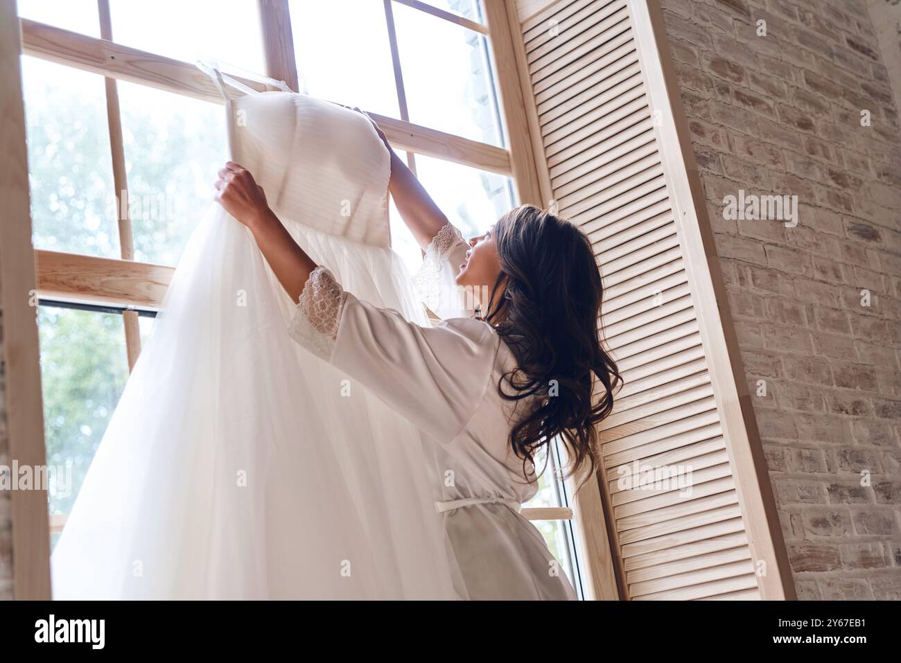 Ich stelle sicher, dass es perfekt ist. Schöne junge Frau im Seidenbademantel, die ihr Hochzeitskleid berührt, während sie am Fenster steht Stockfoto