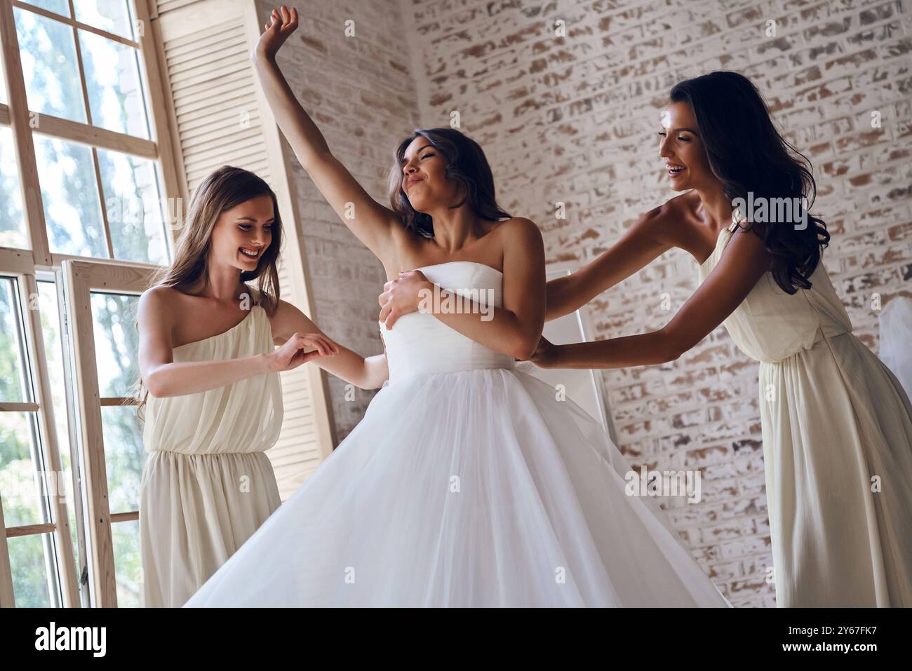 Dafür sorgen, dass alles perfekt ist. Zwei attraktive junge Frauen passen ein Hochzeitskleid an eine schöne Braut an, während sie zusammen am Fenster stehen Stockfoto