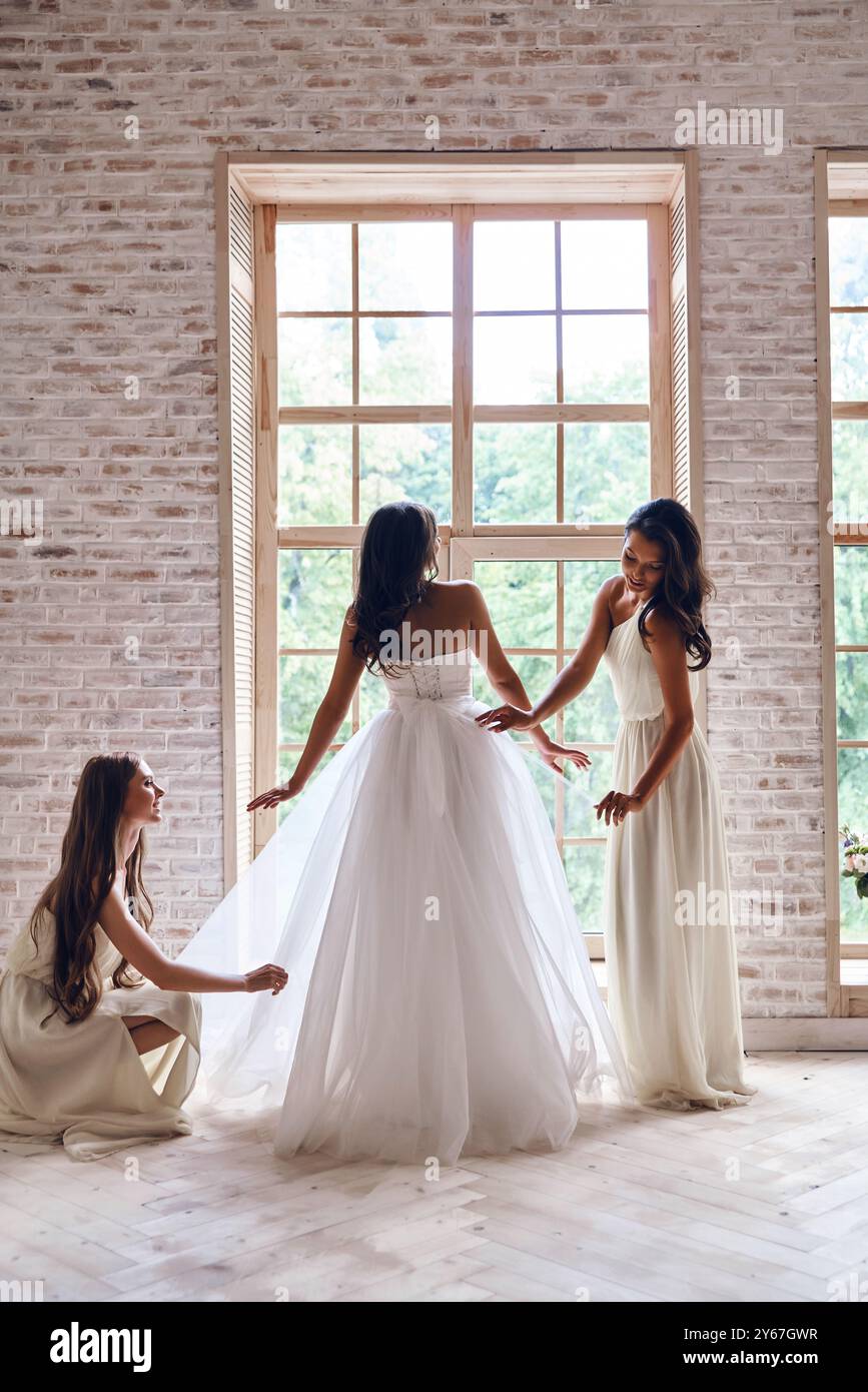 Dafür sorgen, dass sie perfekt aussieht. Die Brautjungfern in voller Länge helfen Braut mit ihrem Hochzeitskleid, während sie zusammen am Fenster stehen Stockfoto