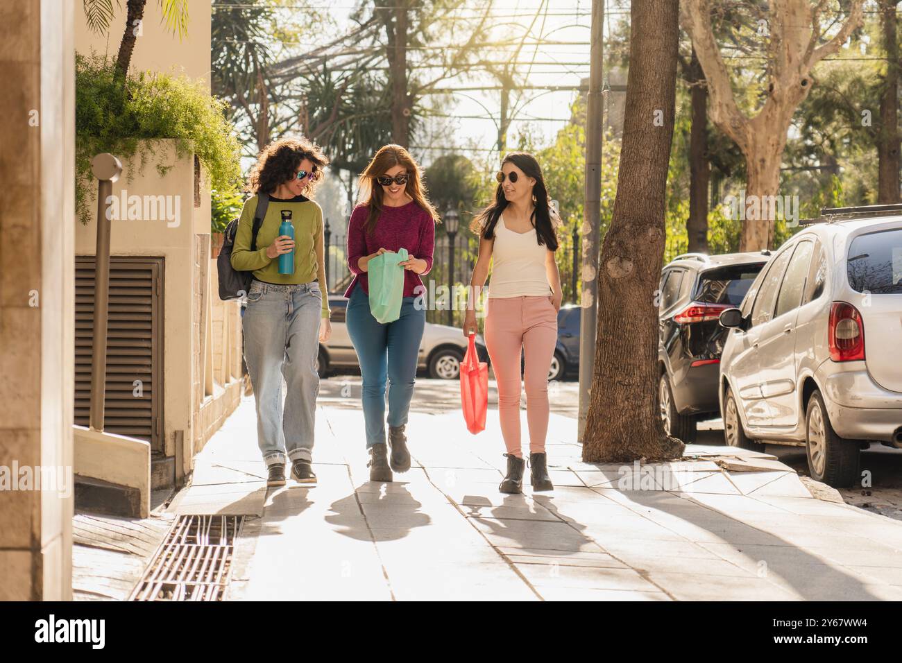 Drei Frauen schlendern auf einem sonnendurchfluteten Bürgersteig, tragen Taschen und unterhalten sich freundlich. Sie sind lässig gekleidet und genießen einen gemütlichen Spaziergang Stockfoto