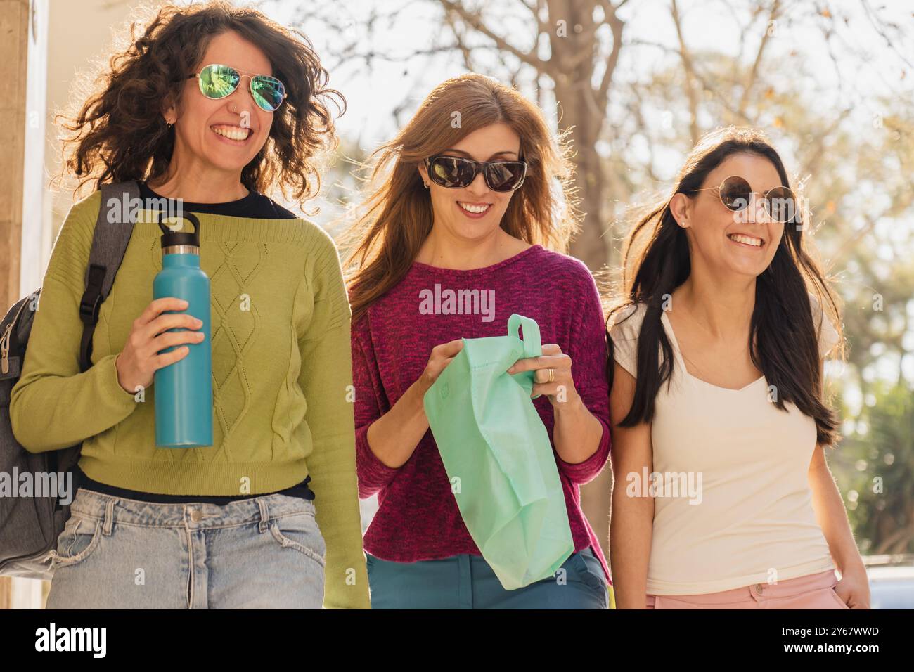 Drei Frauen lächeln und unterhalten sich, während sie draußen im Sonnenlicht laufen. Sie tragen legere Kleidung, Sonnenbrille und tragen umweltfreundliche Taschen und eine Wasserflasche Stockfoto